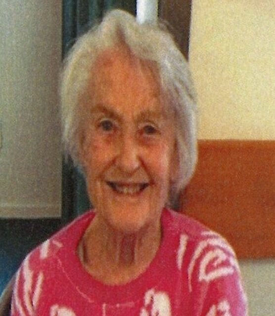 Elderly woman smiling and knitting indoors.