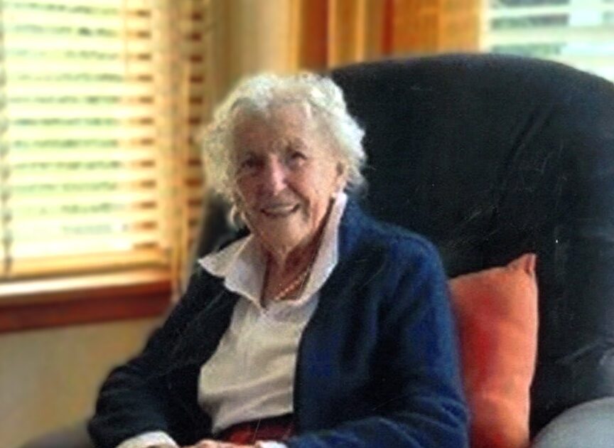 Elderly woman smiling in a tartan skirt.